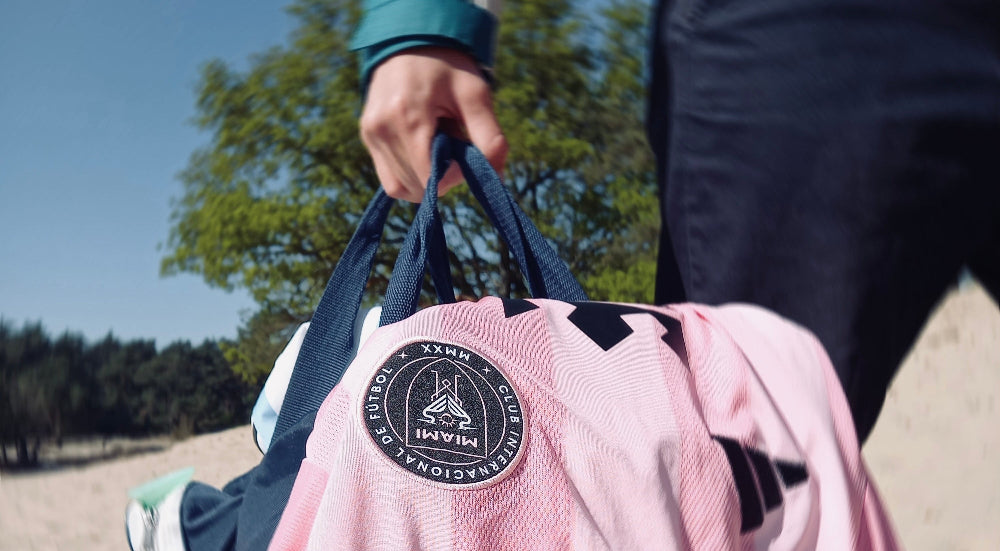 Person holding a pink jacket with a logo and a navy duffel bag on a sandy beach.